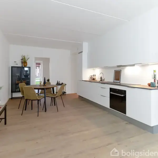 Et moderne køkken-alrum med hvide skabe og integrerede apparater. Spisestuen har et rundt bord med fire stole. En grøn plante står på et lille bord.