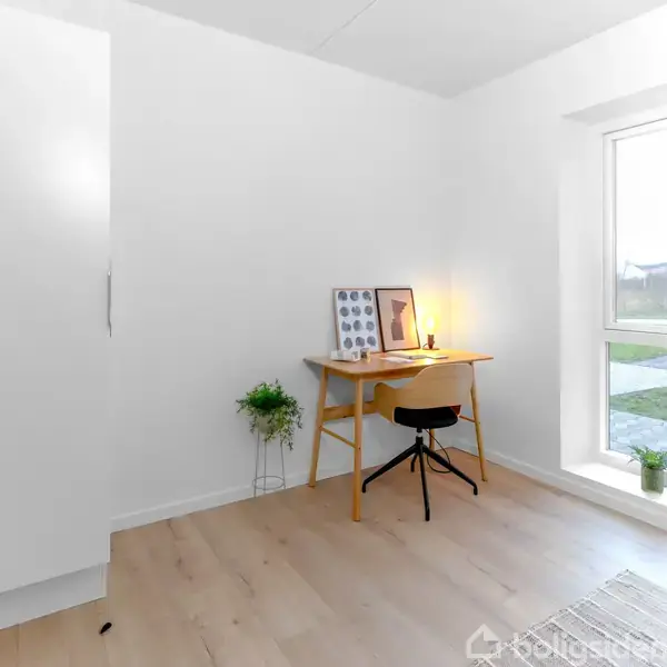 Et minimalistisk kontor med et skrivebord oplyst af en lampe, omkranset af en stol og planter, beliggende i et lyst rum med et stort vindue, udsigt til en græsplæne.