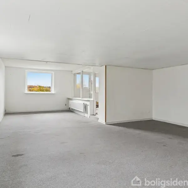 Et tomt rum med gråt gulvtæppe og hvide vægge, indeholder to vinduer, der lukker naturligt lys ind. Rummet er enkelt og rummeligt, ideelt til diverse formål.