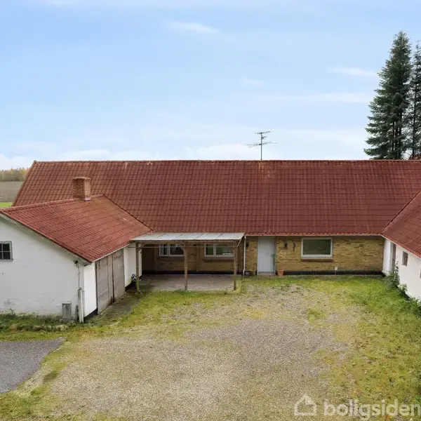 Et hvidt landhus med rødt tegltag står i en gårdsplads, omgivet af grønt græs og en gruppe træer i baggrunden under en blå himmel. Fire sort affaldscontaiere er placeret ved siden.