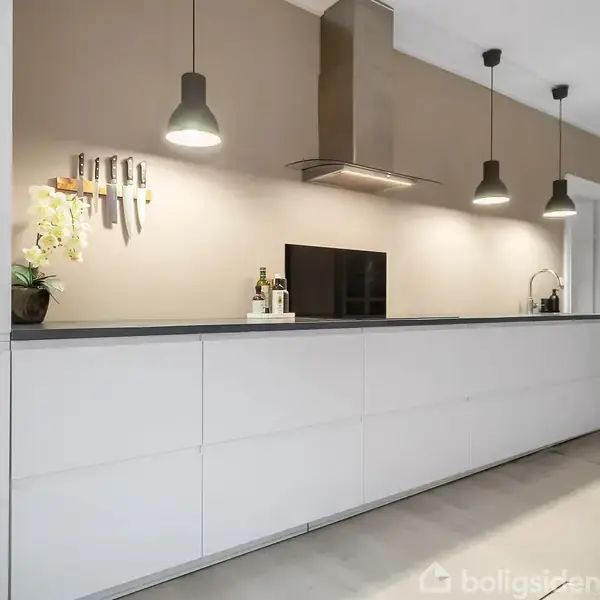 En moderne, lang køkkenbord med hvid front og sort bordplade. Knive hænger på væggen. Tre pendellamper og emhætte hænger over bordet i et stilfuldt køkken.