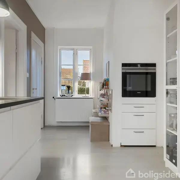 Et moderne køkken med hvidt interiør, hvor en indbygget ovn er placeret ved siden af et glas-displaykabinet fyldt med køkkenudstyr. En vask står til venstre, mens et vindue i baggrunden lukker naturligt lys ind.