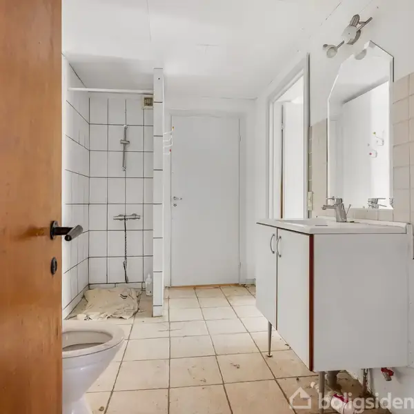 Toilet stående ved siden af en åben dør. Spejl og håndvask placeret på højre side. Flisebelagt bruseområde i baggrunden. Beige flisegulv.