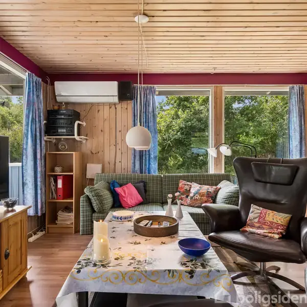 Stue med lænestol og pejs, omgivet af store vinduer med udsigt til grønne træer. Retro sofa, tv på skænk og lille spisebord med dug i midten. Hyggelig atmosfære.