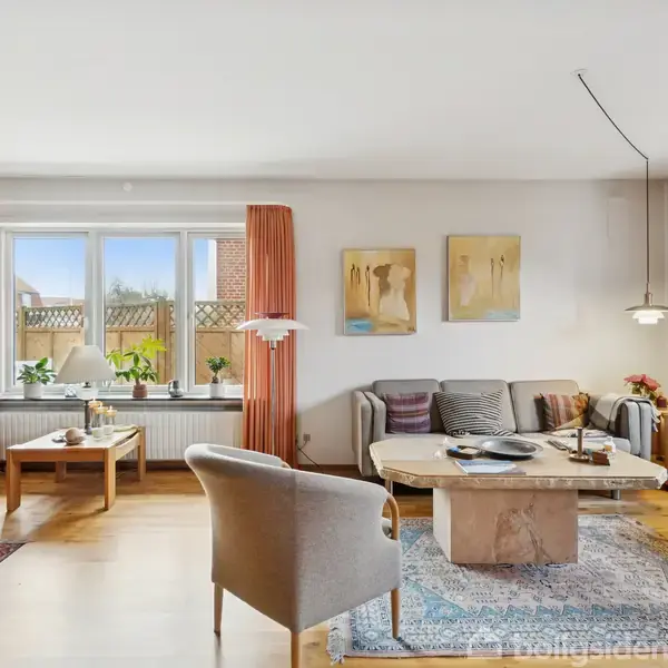 En stue med sofa og stole omkring et sofabord. Rummet har lyse vægge, træmøbler, gulvtæpper, og vinduer med gardiner. Billeder hænger på væggen. Planter står ved vinduet.