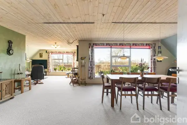 En spisebordgruppe med seks stole står i en lys stue med trælofter, store vinduer og udsigt til haveelementer. Et opholdsområde med lænestol og planter er i venstre side.