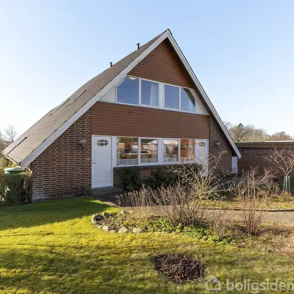 Et hus står stille i en have med græsplæne og buske. Huset har en stejl tagkonstruktion og store vinduer på gavlen. Det er omgivet af træer i baggrunden.