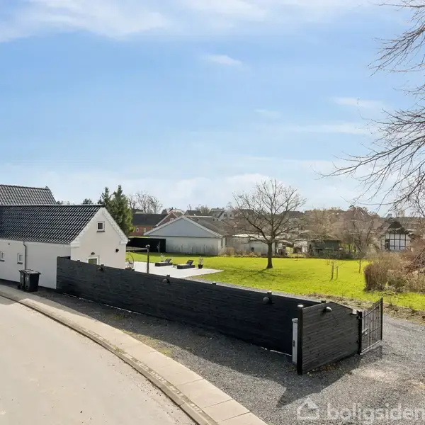 En hvid villa med sort tag står stille ved en asfalteret vej. Omringet af et stort græsareal med enkelte træer, under en blå himmel.