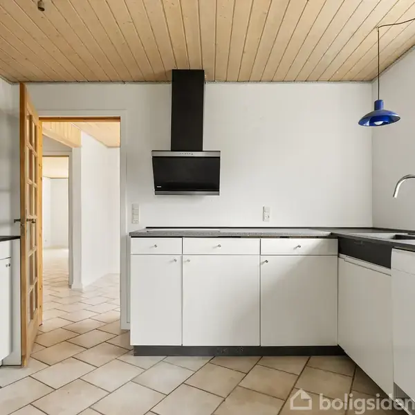 Et moderne køkken med hvide skabe og en sort emhætte. Loftet har træpaneler, og gulvet er flisebelagt. En blå lampe hænger over vasken ved vinduet.