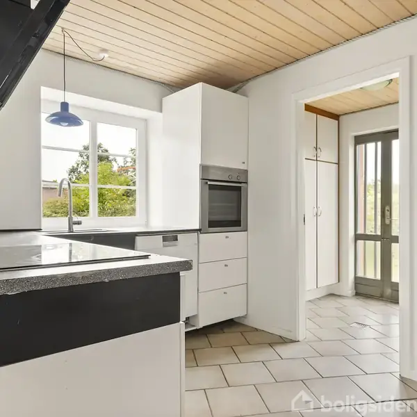 Køkken med hvide skabe og sorte bordplader, kogeplade og indbygget ovn. Der er et vindue med udsigt til træer og en tilstødende entré med glasdør. Ingen tekst.