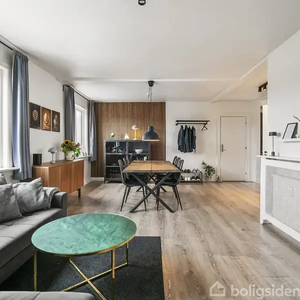 Sofa står foran store vinduer i en moderne stue. Til højre er en spiseplads med et bord og stole, omgivet af hvidt og træindretning. Tekst: "Til salg".