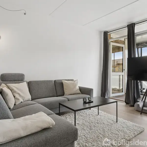 Grå sofa med puder står i hjørnet af en stue. Et sort sofabord står foran den. Der er en stor fladskærm på et stativ. Gardiner hænger ved en altandør.