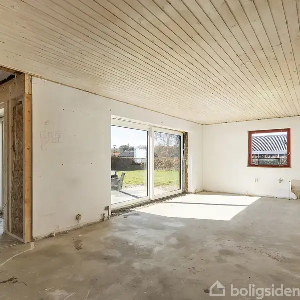Rum under renovering med rå cementgulv og hvidmalede vægge. Store skydedøre leder ud til en have. Træloftet er ubehandlet, og et rødt vindue ses til højre.