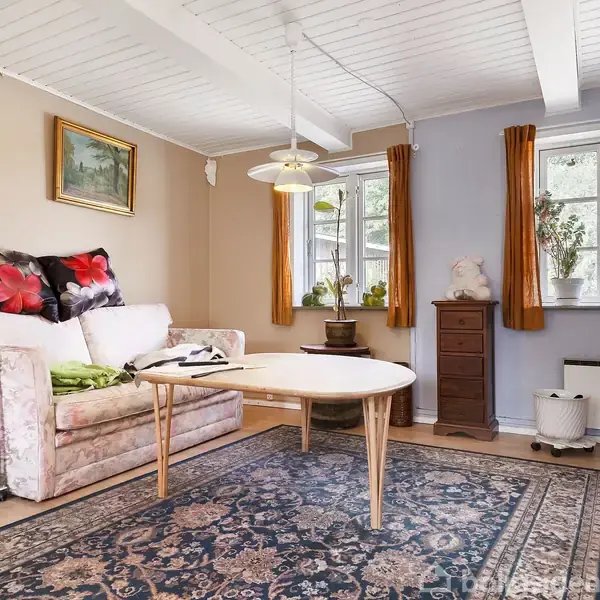 Sofa med blomstrede puder står stille ved væggen i en stue. I rummet er et rundt træbord, en orientalsk tæppe, og vinduer med orange gardiner. En kommode står til højre.