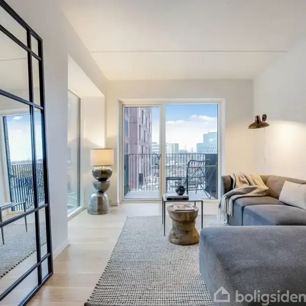 Stue med grå hjørnesofa, sofabord, og tæppe. Store vinduer viser en balkon med udsigt over byen. Højt spejl til venstre giver rummelig fornemmelse. Lampeskærm ved sofaen.
