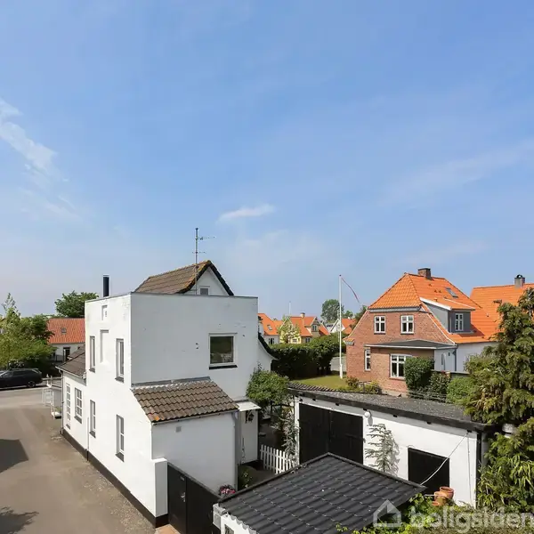 Hvidt hus med fladt tag står stille i et villakvarter med andre traditionelle murstenshuse. Solen skinner, og himlen er let skyet. Ingen tekst findes i billedet.