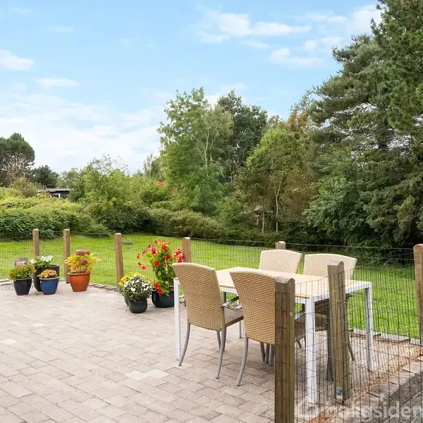 Terrassemøbler står på en flisebelagt terrasse. Fire stole omgiver et bord. Flere potteplanter er placeret omkring, mens grønne træer og buske danner baggrunden under en blå himmel.