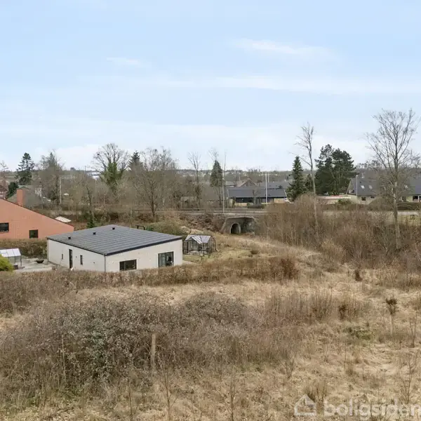 En række huse omkranser et åbent, græsbevokset område med nøgne træer og buske. Bygningerne er spredt, giver en fornemmelse af et landligt boligområde.