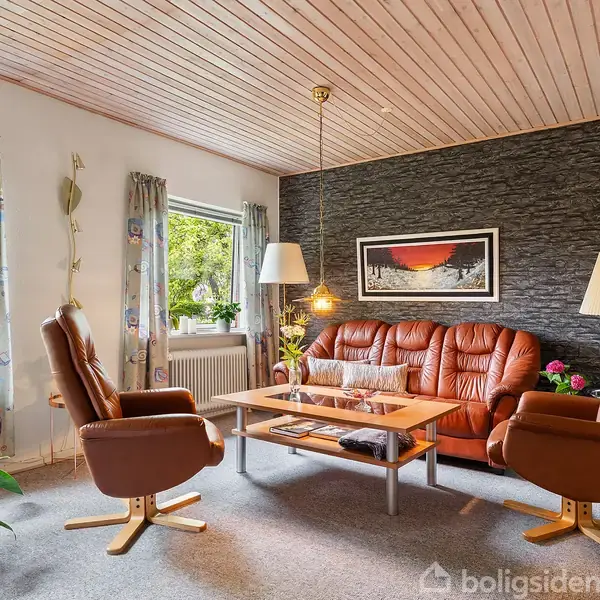 Stue med brunt lædermøblement, hvor en sofa og to lænestole omgiver et sofabord. Væggene har en sten-tekstur, mens lamper og grønne planter tilføjer hygge. Maleri hænger på væggen.