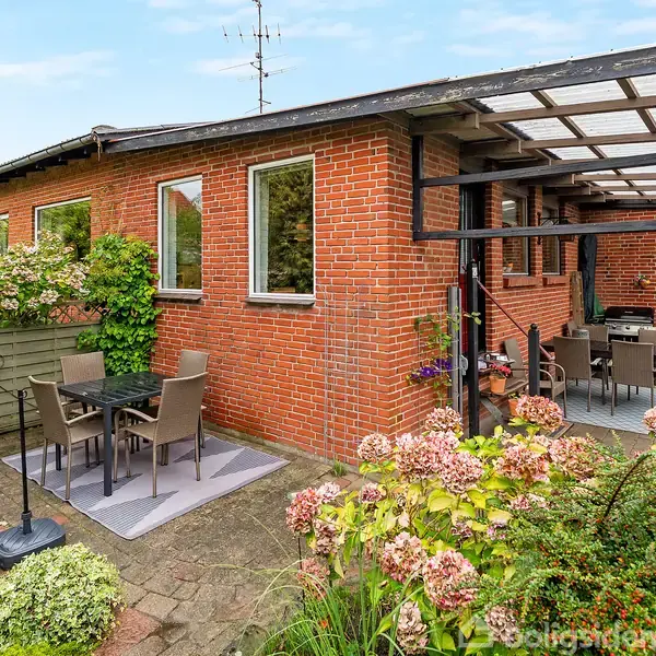 Murstenshus med terrasse. Haven indeholder bord og stole til afslapning. Planter og blomster omringer området. En overdækket del mod huset har flere stole, hvilket skaber en hyggelig atmosfære.