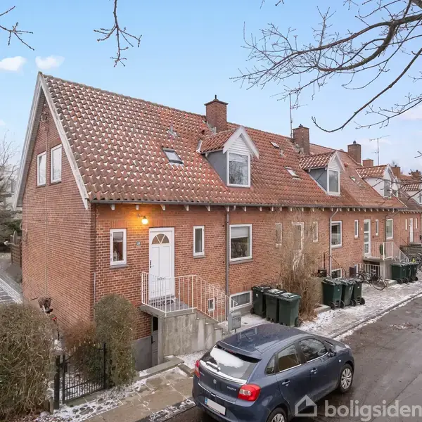 Rækkehus i røde mursten står på række langs en snedækket vej, hvor biler er parkeret. Huset har et rødt tegltag og flere kviste. Ingen synlig tekst.