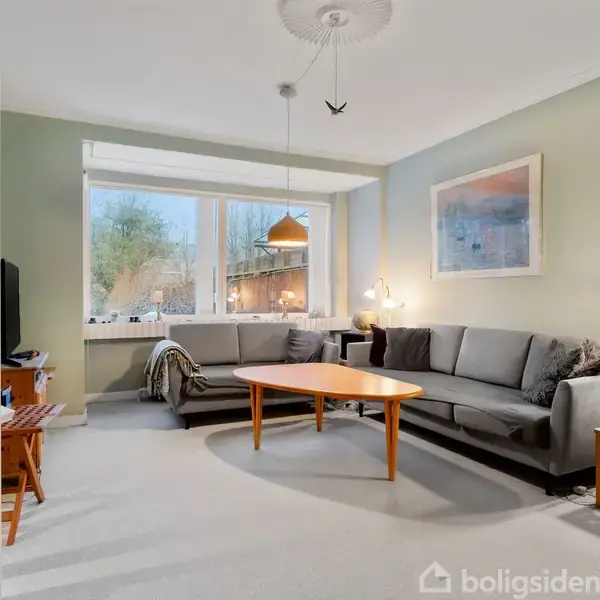 En stue med en grå hjørnesofa og et rundt træbord i midten. Rumoplyst af naturligt lys fra et stort vindue. Der hænger lamper og billeder på væggene.