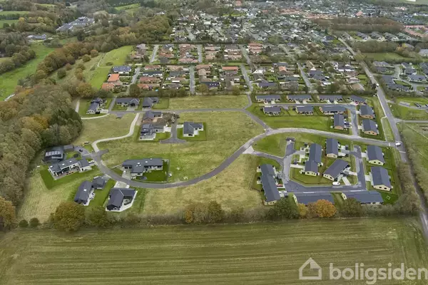 Luftfoto af et boligområde med moderne huse, placeret omkring snoede veje. Græsarealer omgiver husene, mens en større bydel ses i baggrunden med tættere bebyggelse.