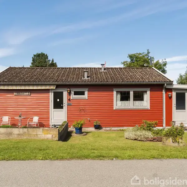 Rødt hus står stille med hvide vinduer og døre. Foran er der en grøn græsplæne med blomsterbede og en indkørsel. Et skilt viser: "Piberbakken 5".
