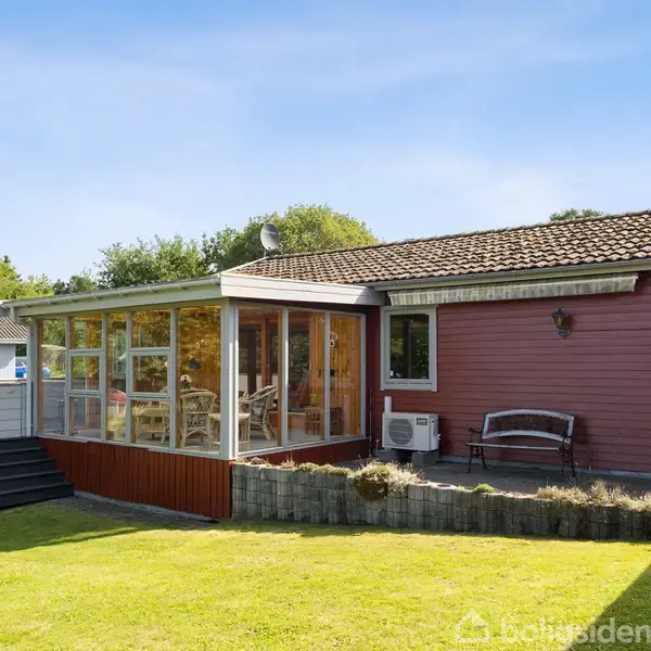 Et rødt træhus med en stor, indglasset veranda fyldt med møbler. Huset er omgivet af en velplejet græsplæne og buske, med en bænksæt langs husets ydre mur.