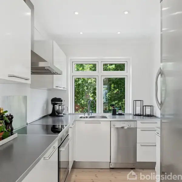 Et moderne køkken med hvide skabe og en grå bordplade har en inklineret opvaskemaskine og en stor køleskab. En skål frugt står ved siden af en håndmikser. Vindue viser grønne træer udenfor.