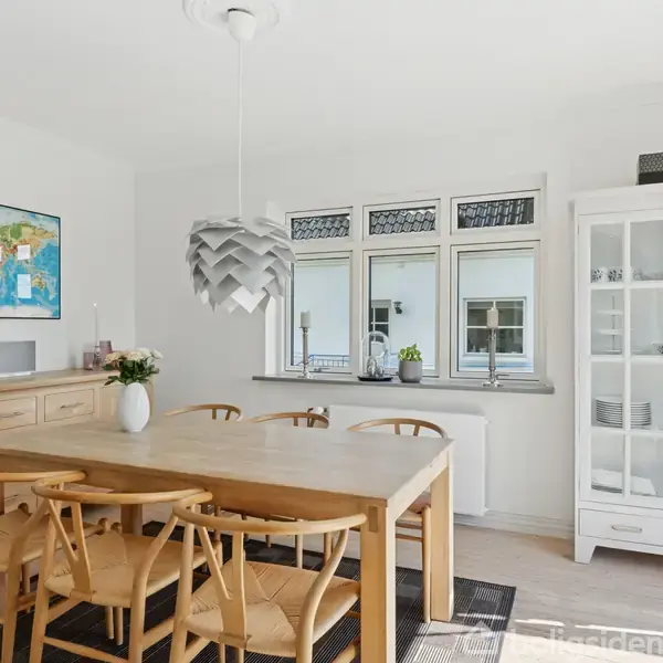 Spisebord med træstole står i et lyst køkken/alrum. En moderne lampe hænger over bordet. Til venstre ses et skab og verdenskort, og tre vinduer i baggrunden.