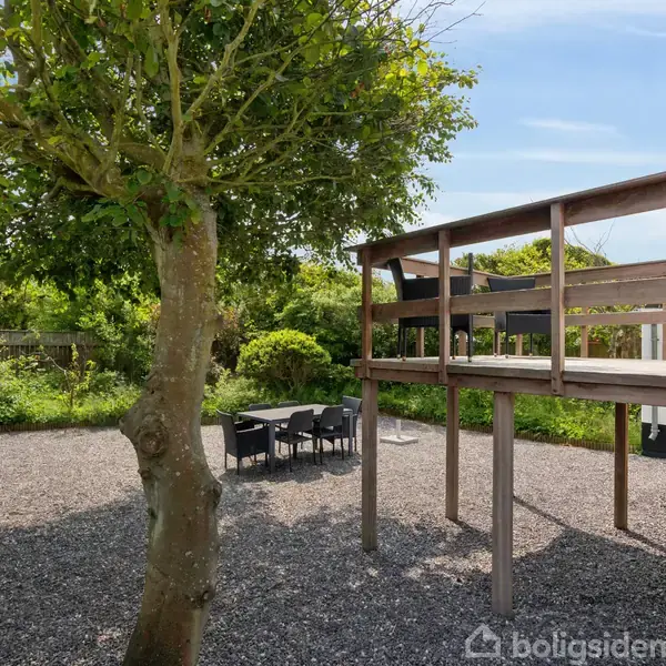 En træterrasse står forhøjet, med trappeadgang, ved siden af et havemiljø med stenbelægning og et træ. Et havebord med stole står under et stort skyggefuldt træ.