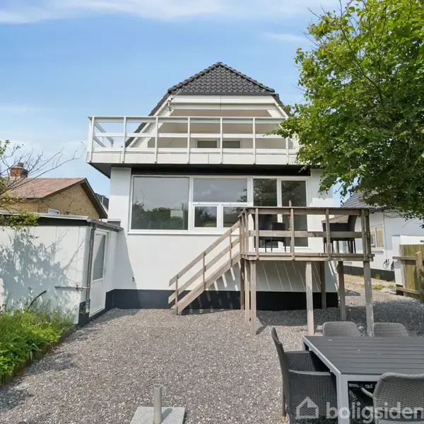 Et to-etagers hus med en balkon øverst. En trappe leder til indgangen på første sal. Foran huset er der en terrasse med et spisebord i en haveomgivelser.
