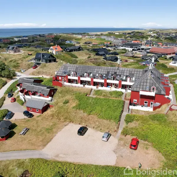 Rød bygning med røde tage ligger på en grøn bakke, omgivet af små veje og sommerhuse. Flere biler er parkeret omkring bygningen. I baggrunden ses klitter og havet.