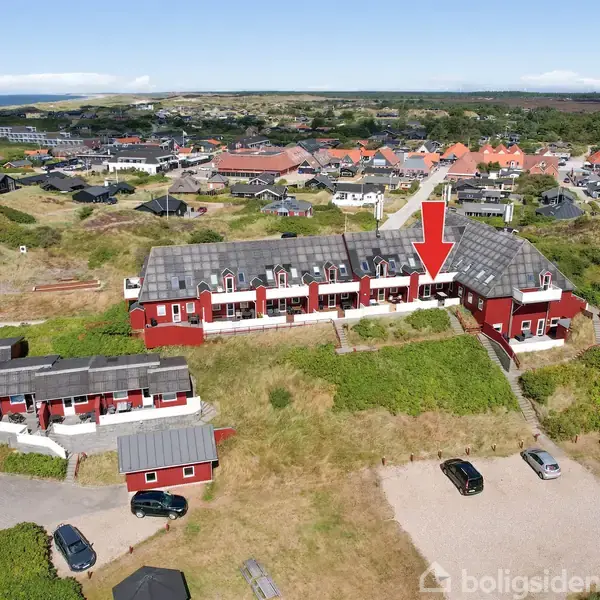En række røde bygninger med hvide vinduer set fra oven, placeret i et landligt boligområde. En stor rød pil peger mod én af bygningerne. Flere biler er parkeret i nærheden.