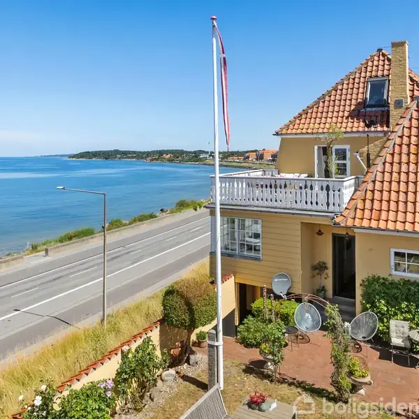 Et gult hus med rødt tegltag har en balkon med udsigt over havet. Det ligger ved en vej, omgivet af have. Flagstang rejser sig i forgrunden.