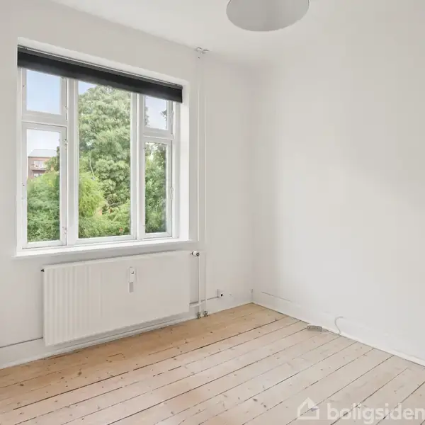 Et tomt rum indeholder et stilrent trægulv uden møbler. Et stort, åbent vindue med hvid karm giver udsigt til grønne træer udenfor, og en radiator er placeret under vinduet.