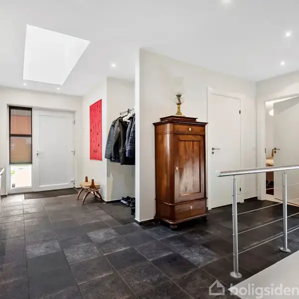 Garderobe i gangareal med skiferfliser, tøj på knagerække, et mørkt træmøbel, og et rundt spejl på væggen; døre fører til andre rum. Ingen tekst fundet.