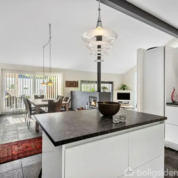 Et moderne køkken-alrum med en sort bordplade, skuffer og skabe. Der ses en spiseplads i baggrunden med stole, et rødt gulvtæppe, og en stue med lysindfald fra vinduer.