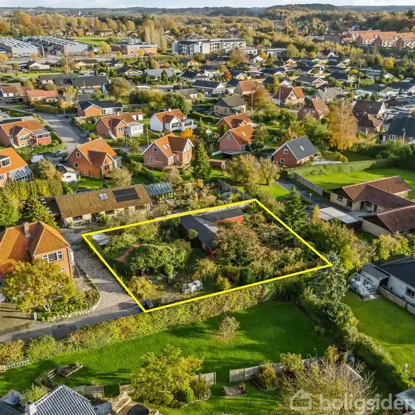 En ejendom omgivet af en gul markering, dækker en have med træer og buske i et parcelhuskvarter med flere huse. Det grønne landskab breder sig over et stort område.