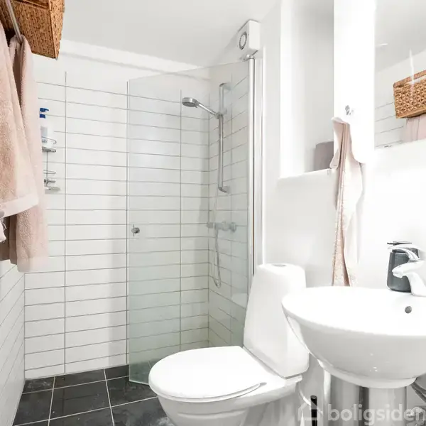 Toilet er placeret ved siden af en vask med spejl, mens brusekabinen med glasvæg befinder sig for enden af det smalle badeværelse. Hvide fliser og sort gulvbelægning dominerer rummet.