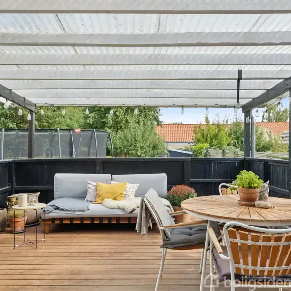 Grill og havemøbler står på en overdækket terrasse med trægulv. Sofaen har puder, og rundt bord med stole står ved siden af planter. Udsigt til grønne buske og nabotag.