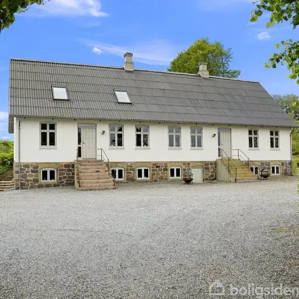 En hvid murstensbygning med sort tag og flere vinduer står stille. Den er omgivet af grus, grønne buske, og en skyfri blå himmel.