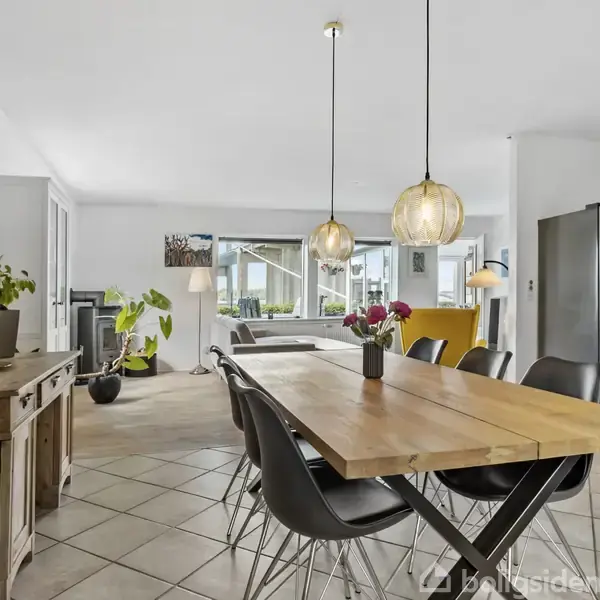 Langt spisebord i træ med sorte stole omkring står i et rummeligt køkken-alrum. Over bordet hænger to pendellamper. Baggrunden indeholder sofaer, kunstværk og planter.