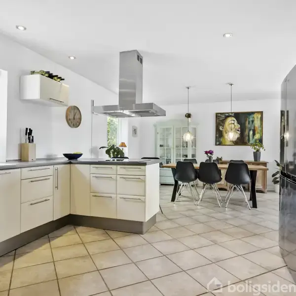 Køkken med hvide skabe og stålapparater; åbent mod spiseområde med sorte stole; store vinduer slipper naturligt lys ind; grønne planter pryder rummet; maleri og ur på væggen.