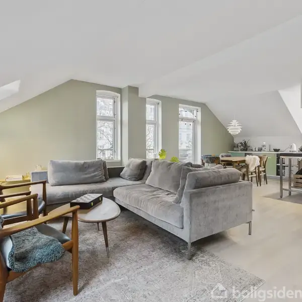 Grå sofaarrangement med træstole står på et tæppe i en lys stue. I baggrunden er et spisebord og et moderne køkken med øer under skrå lofter og ovenlysvinduer.