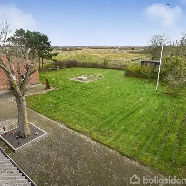 Et grønt græsareal ligger ved siden af en rød murstensbygning. Et flagstang står på plænen, omgivet af hække og åbne marker i baggrunden. En brolagt terrasse omkranser et træ.