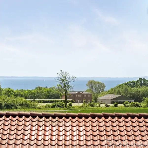 Rødt tegltag ses gennem et vindue, med et landskab i baggrunden bestående af grønne træer, en vej og fjern udsigt over havet under en klar blå himmel.