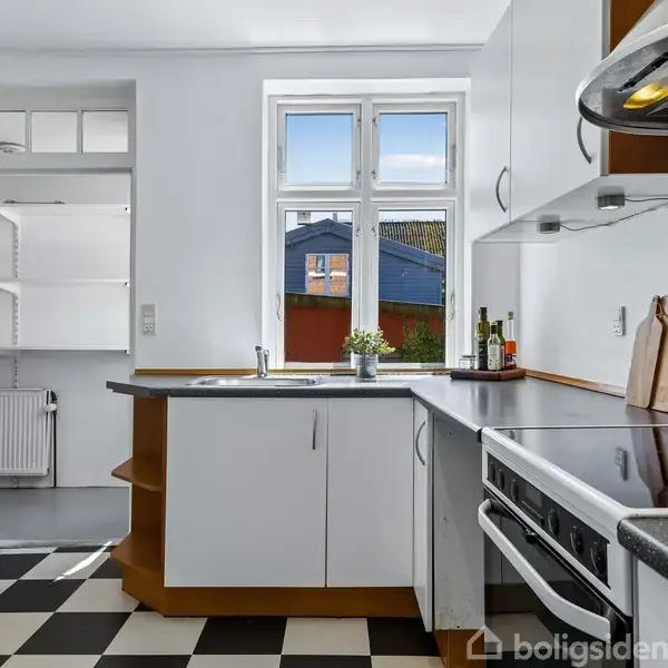 Et køkken med sorte og hvide ternede gulve. Hvide skabe står rundt om en sort bordplade med en vask. Et vindue med udsigt til et blåt hus bagved.