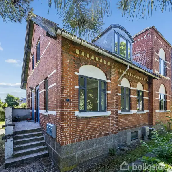 Stue med sofa og stole står overfor et glasbord, omgivet af grønne planter. Spisebord til seks personer placeret ved vinduet. Trægulv og loft giver en hyggelig atmosfære.